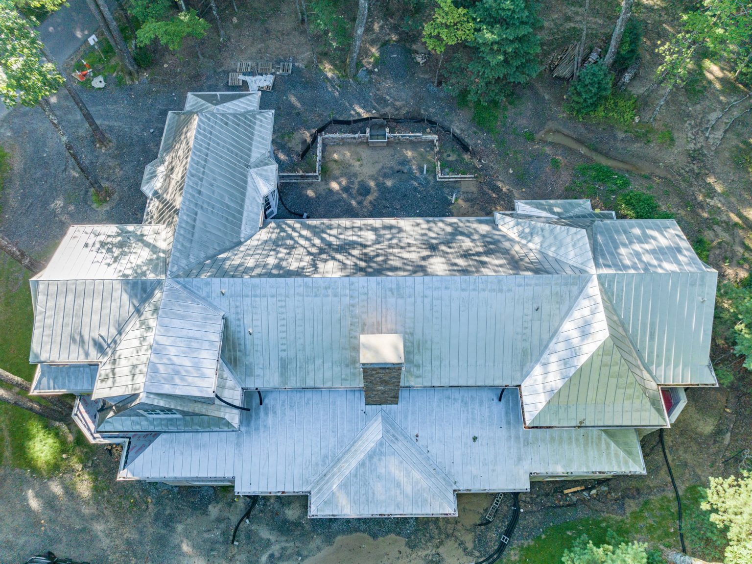 Aerial view of a large residential house under construction, featuring an extensive, light gray standing seam metal roof, a stone chimney, and dirt landscaping.