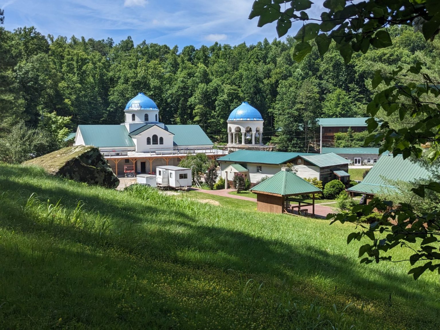 Exterior view of the Holy Cross Monastery church and surrounding buildings in Wayne, West Virginia, featuring white stucco walls, blue metal domes, and green metal pitched roofs set against a forested hillside.