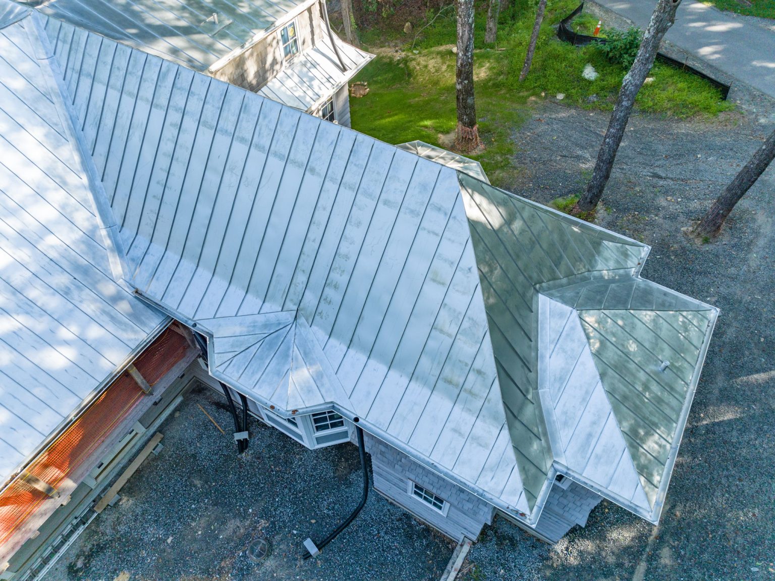An aerial photograph of a building with a complex, multi-gabled, light gray standing seam metal roof installation, showing various angles and roof planes.