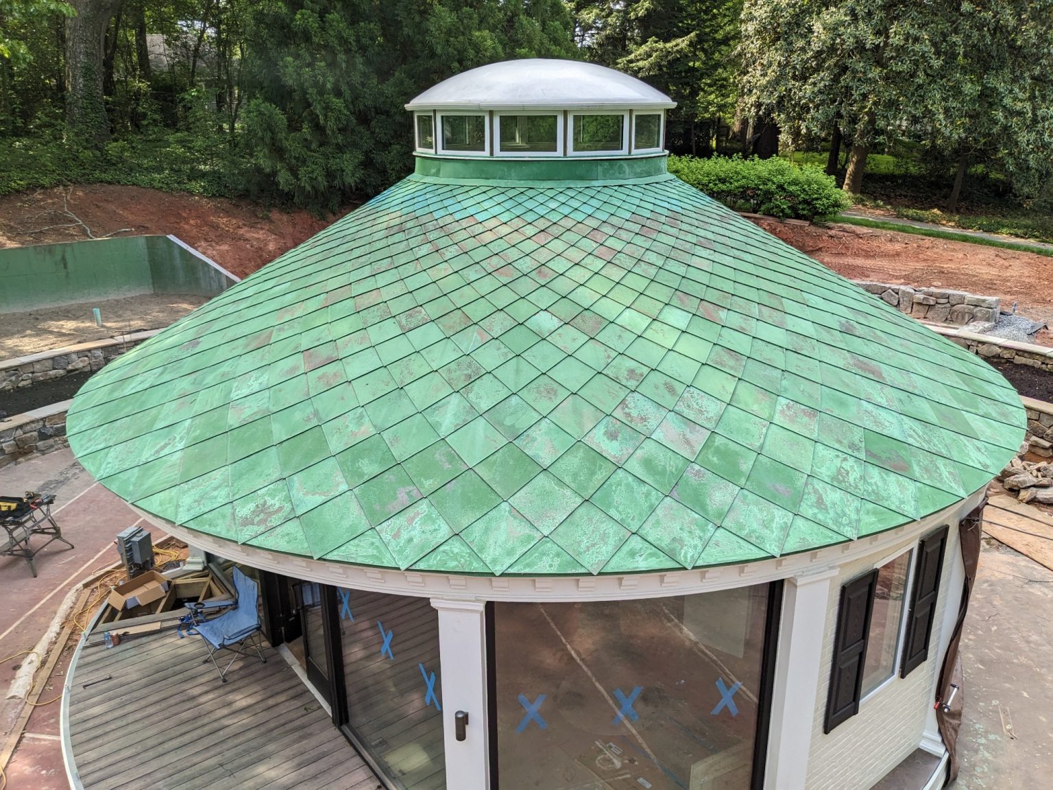 A single-story, light-yellow, curved garden structure featuring a green metal roof with diamond-pattern shingles and a small cupola, set within a lush green lawn and surrounded by deciduous trees with autumn foliage.