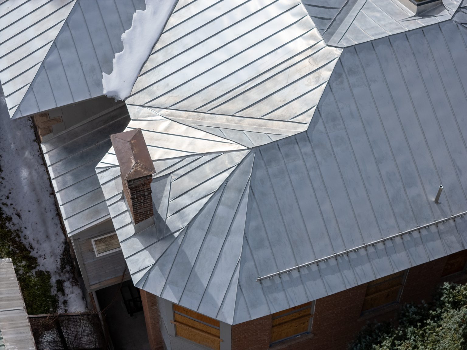 An overhead view of the complex, multi-pitched standing seam metal roof of the Ouray County Museum, showing the various roof planes and flashing details around a brick chimney, with a small patch of snow visible near the eave.