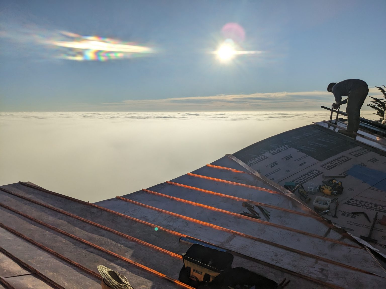 A roofer works on the installation of a standing seam copper roof high above a dense layer of white clouds at sunset.