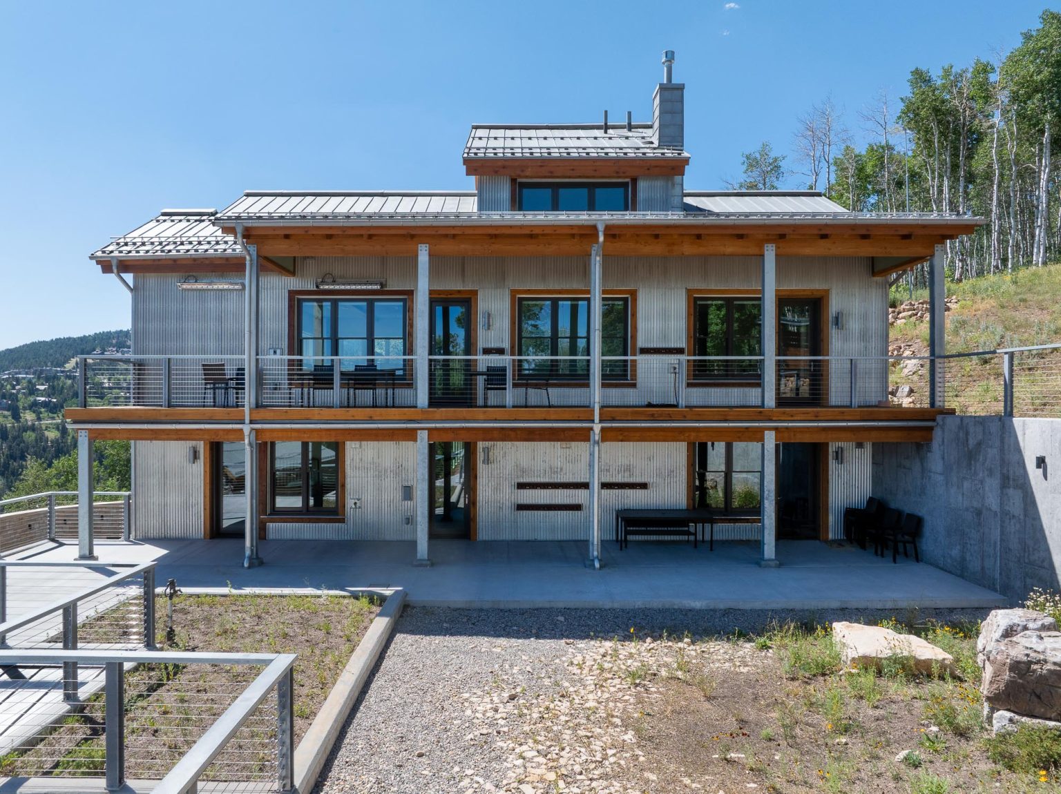 A two-story modern mountain house with a gray standing seam metal roof, wooden siding, two levels of decks with cable railings, and a concrete foundation built into a hillside landscape.